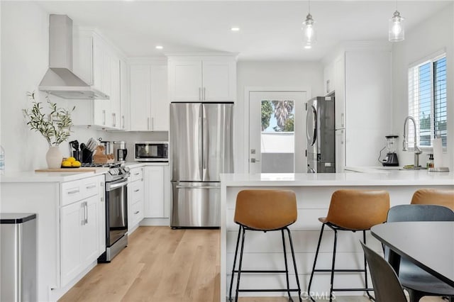 kitchen featuring light wood-type flooring, stainless steel appliances, wall chimney range hood, decorative light fixtures, and white cabinetry