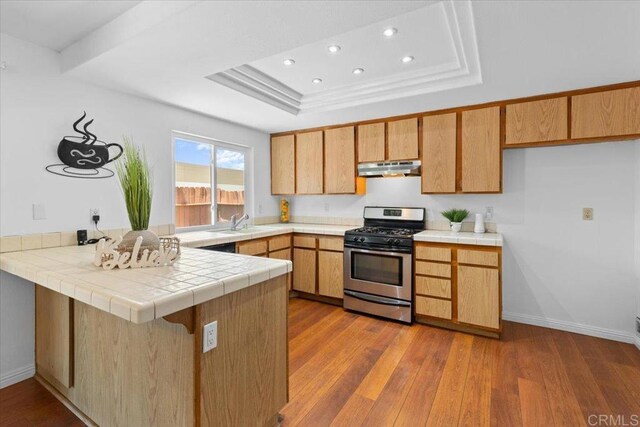 kitchen with gas stove, a raised ceiling, kitchen peninsula, tile countertops, and light hardwood / wood-style floors