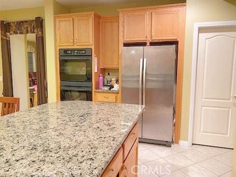 kitchen featuring light brown cabinetry, light stone counters, black double oven, stainless steel refrigerator, and light tile patterned flooring