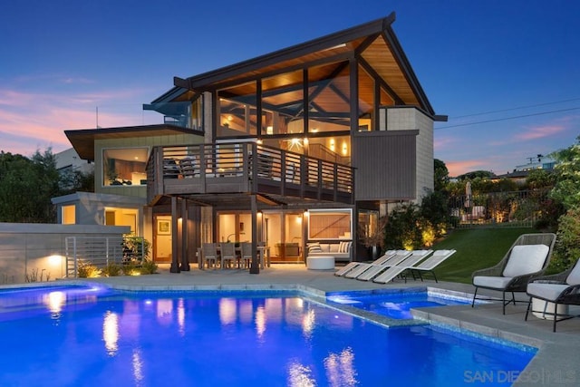 back house at dusk featuring a swimming pool with hot tub, a balcony, and a patio