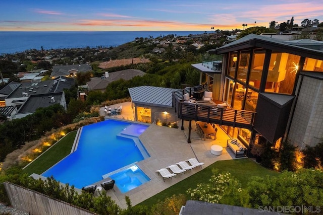 pool at dusk featuring a patio, a water view, and a lawn