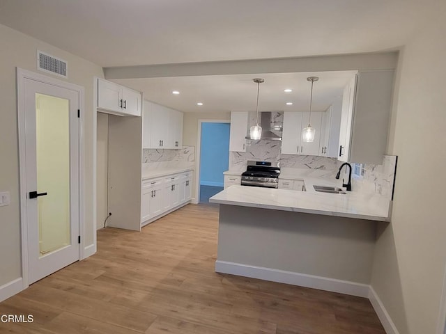 kitchen featuring stainless steel gas stove, sink, wall chimney range hood, decorative light fixtures, and white cabinets