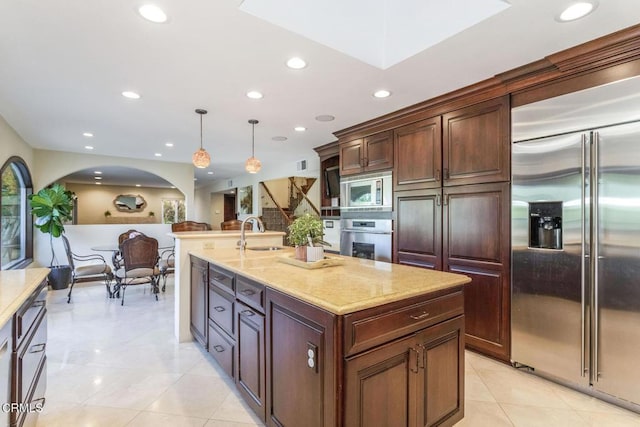 kitchen featuring sink, a kitchen island with sink, built in appliances, light stone countertops, and decorative light fixtures