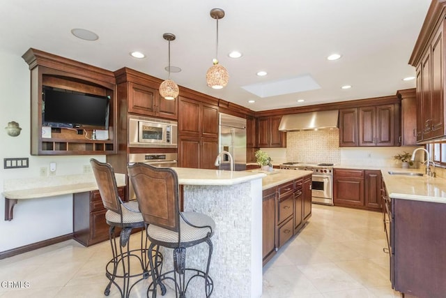 kitchen with pendant lighting, sink, wall chimney range hood, built in appliances, and a center island with sink