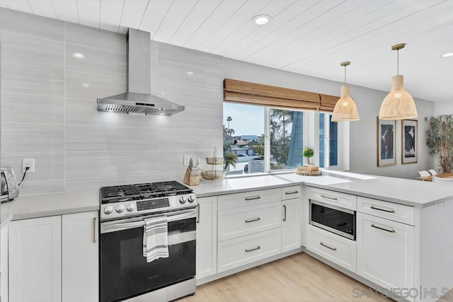 kitchen featuring wall chimney range hood, appliances with stainless steel finishes, decorative light fixtures, white cabinets, and light wood-type flooring