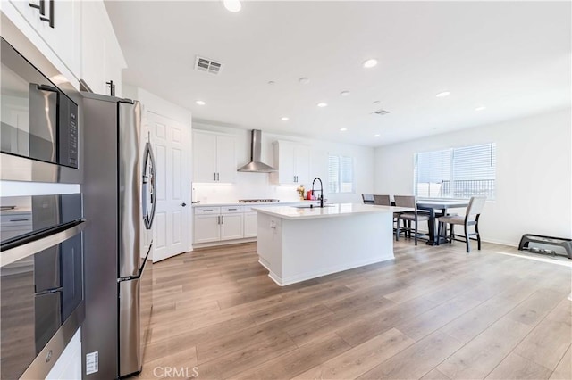 kitchen featuring appliances with stainless steel finishes, wall chimney range hood, a center island with sink, light hardwood / wood-style flooring, and white cabinets