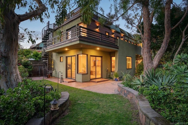 back house at dusk featuring a lawn, a patio, and a balcony