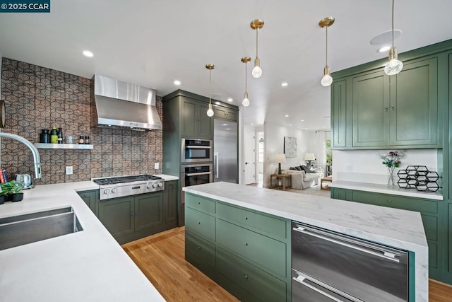 kitchen featuring stainless steel appliances, wall chimney range hood, decorative light fixtures, green cabinetry, and sink