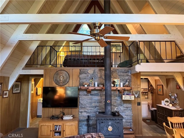 living room featuring beam ceiling, a wood stove, ceiling fan, high vaulted ceiling, and wood walls