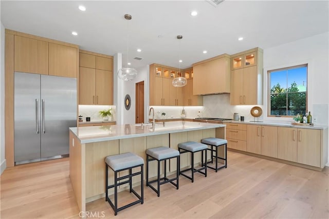 kitchen with custom exhaust hood, stainless steel appliances, light brown cabinets, light hardwood / wood-style flooring, and an island with sink