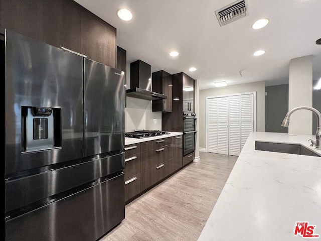 kitchen featuring sink, wall chimney range hood, light stone counters, light hardwood / wood-style floors, and appliances with stainless steel finishes