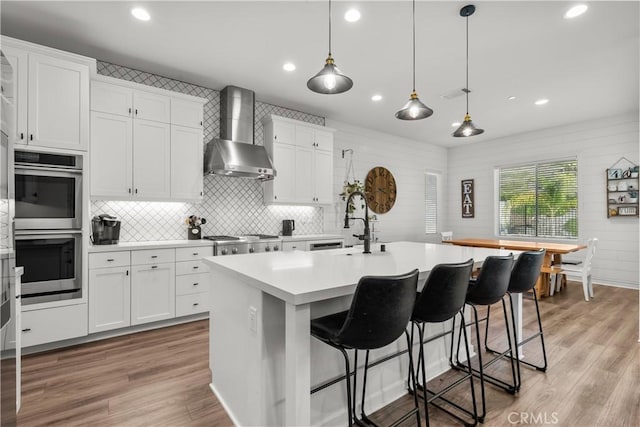 kitchen featuring a center island with sink, wall chimney exhaust hood, light wood-type flooring, and white cabinetry