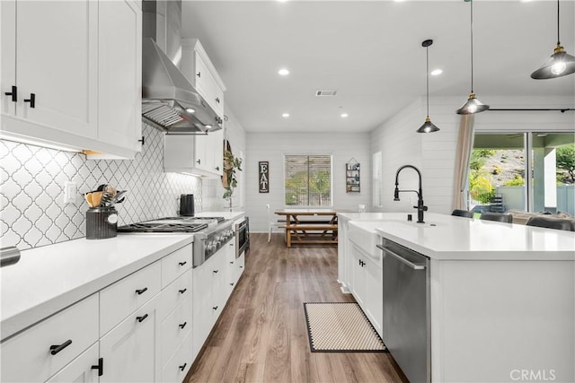 kitchen with appliances with stainless steel finishes, decorative light fixtures, wall chimney exhaust hood, and an island with sink