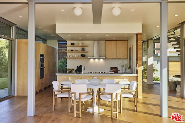 kitchen featuring light brown cabinets, wall chimney range hood, a wall of windows, black double oven, and light hardwood / wood-style floors