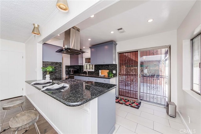 kitchen featuring tasteful backsplash, kitchen peninsula, dark stone countertops, island exhaust hood, and a breakfast bar area