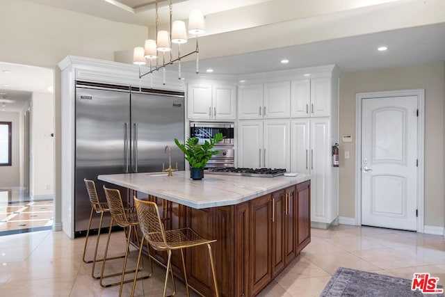 kitchen featuring white cabinetry, built in appliances, an island with sink, pendant lighting, and light tile patterned flooring