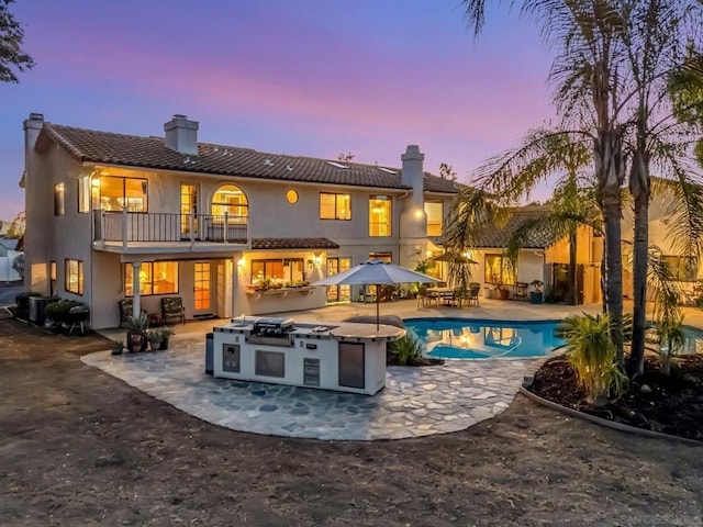 back house at dusk featuring a balcony, exterior kitchen, and a patio