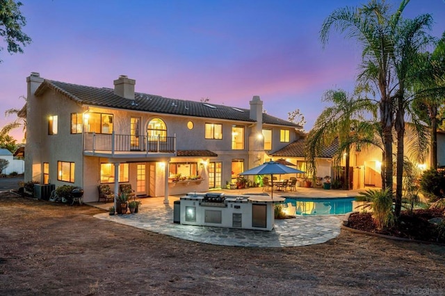 back house at dusk featuring an outdoor kitchen, a patio area, a balcony, and central AC