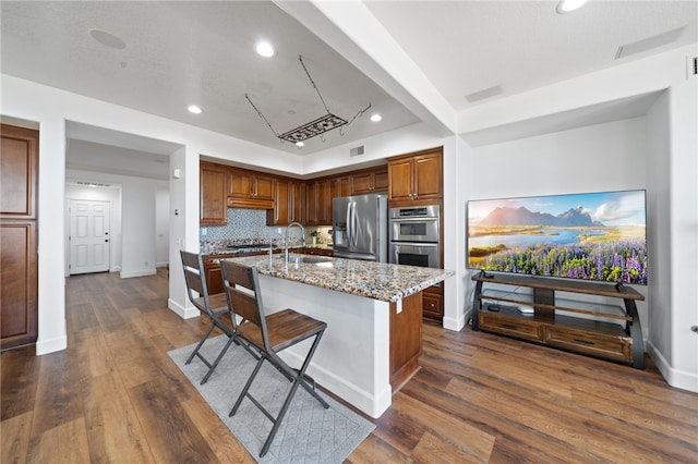 kitchen with backsplash, a kitchen island with sink, appliances with stainless steel finishes, a kitchen breakfast bar, and light stone counters