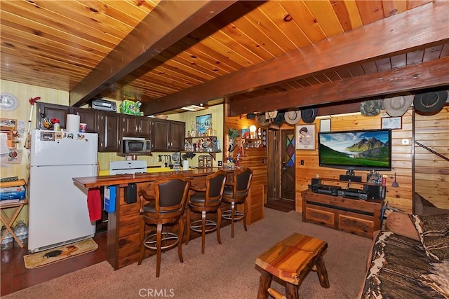 kitchen with wood walls, light carpet, beamed ceiling, white fridge, and dark brown cabinetry