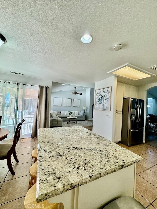 kitchen with light tile patterned floors, white cabinets, a ceiling fan, black fridge with ice dispenser, and light stone countertops