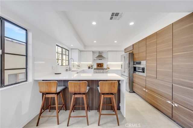 kitchen featuring kitchen peninsula, stainless steel appliances, sink, wall chimney range hood, and white cabinets