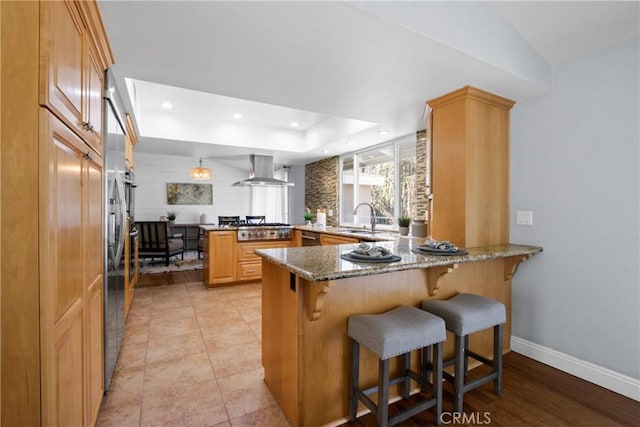 kitchen featuring island range hood, a raised ceiling, dark stone countertops, a peninsula, and a sink