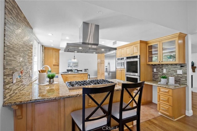 kitchen featuring glass insert cabinets, light stone counters, a peninsula, island exhaust hood, and stainless steel appliances