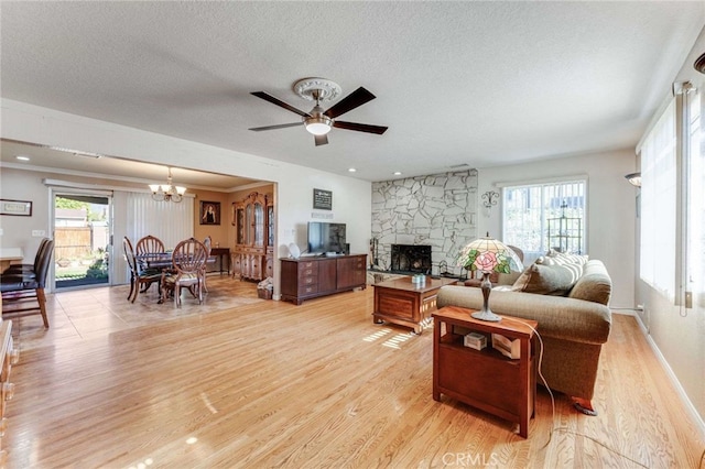 living room featuring light wood-type flooring, a textured ceiling, ceiling fan with notable chandelier, and a stone fireplace