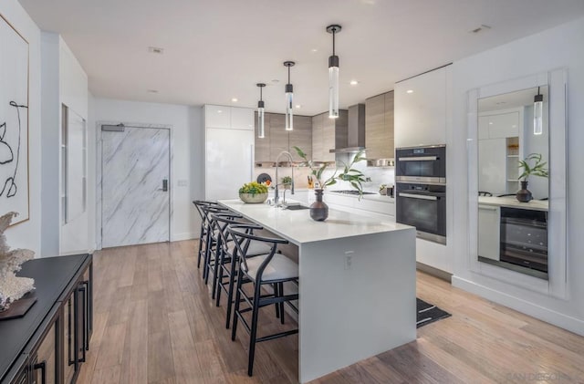 kitchen with pendant lighting, white cabinets, light hardwood / wood-style flooring, wall chimney range hood, and a center island with sink