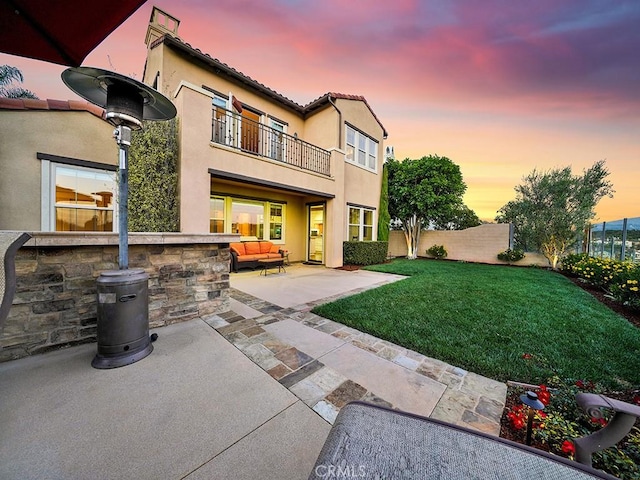 back house at dusk with an outdoor living space, a patio area, a balcony, and a lawn