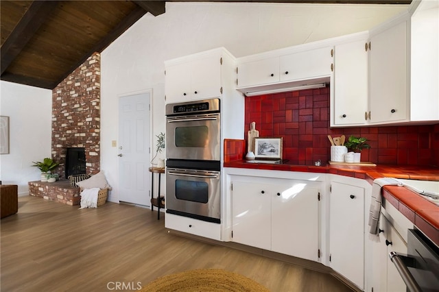 kitchen with a fireplace, white cabinetry, light wood-type flooring, stainless steel double oven, and beam ceiling