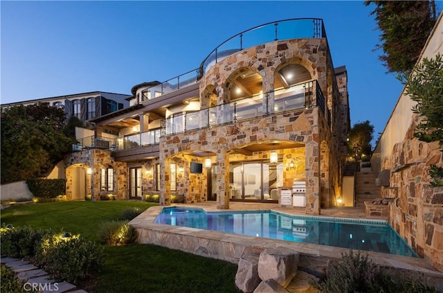 back house at dusk with a balcony, a patio area, and a lawn