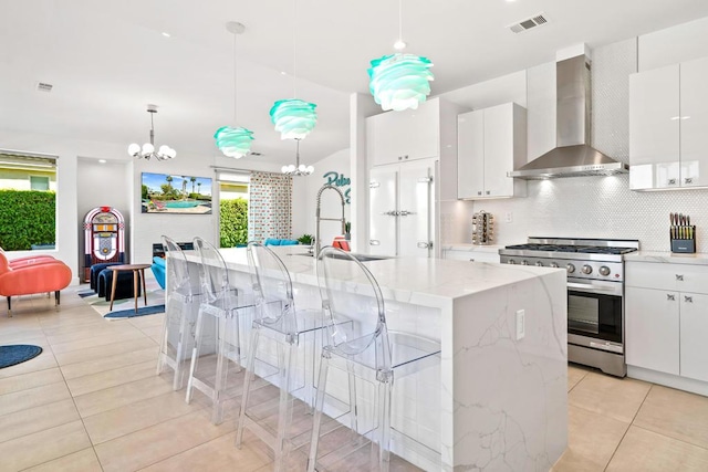 kitchen featuring stainless steel gas range oven, white cabinetry, wall chimney exhaust hood, and an island with sink