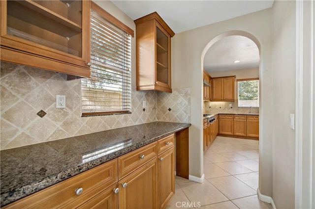 kitchen with light tile patterned floors, arched walkways, dark stone counters, brown cabinetry, and glass insert cabinets