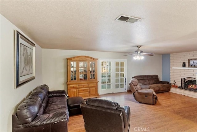 living area with visible vents, light wood-style floors, a brick fireplace, ceiling fan, and a textured ceiling