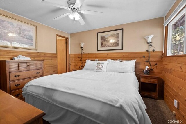 bedroom featuring a wainscoted wall, wood walls, dark carpet, and a ceiling fan