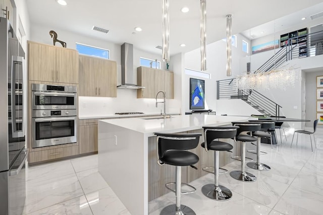 kitchen with sink, stainless steel appliances, wall chimney range hood, pendant lighting, and a kitchen island with sink