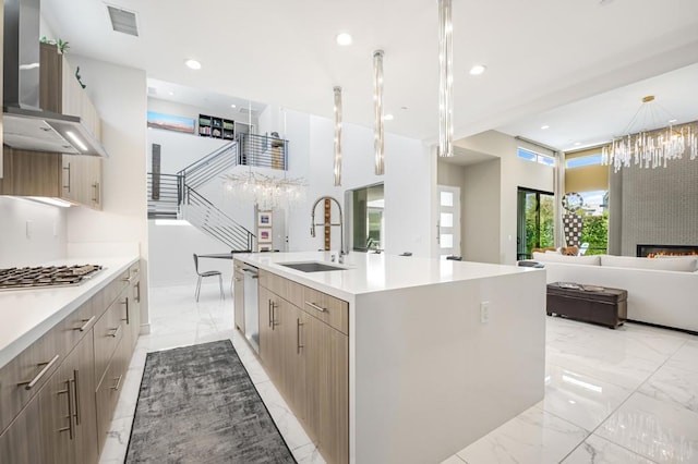 kitchen with stainless steel appliances, a kitchen island with sink, wall chimney range hood, sink, and decorative light fixtures