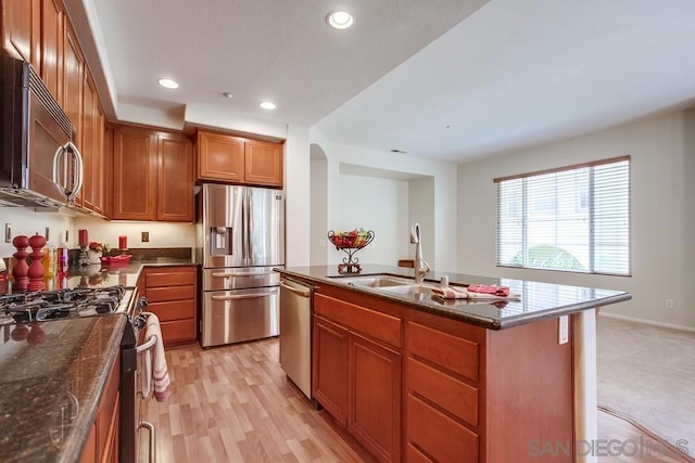 kitchen with appliances with stainless steel finishes, a kitchen island with sink, sink, dark stone countertops, and light hardwood / wood-style floors