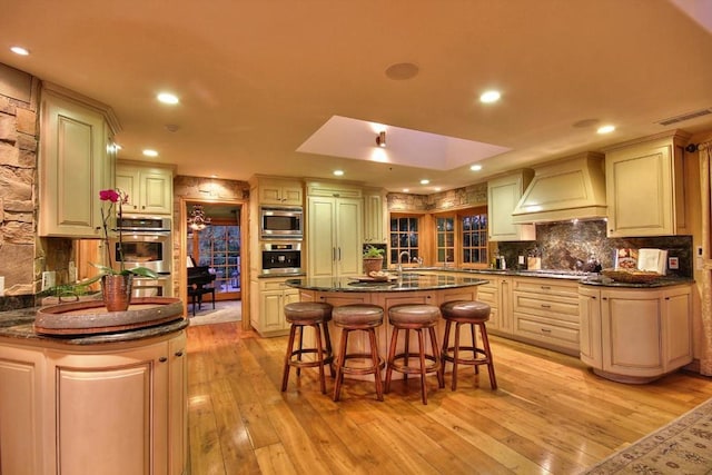 kitchen with a center island with sink, light hardwood / wood-style floors, dark stone countertops, and custom range hood