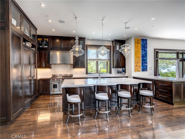 kitchen featuring a kitchen island with sink, wall chimney range hood, decorative light fixtures, range with two ovens, and dark hardwood / wood-style floors