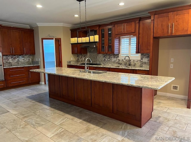 kitchen with decorative backsplash, light stone countertops, sink, hanging light fixtures, and an island with sink