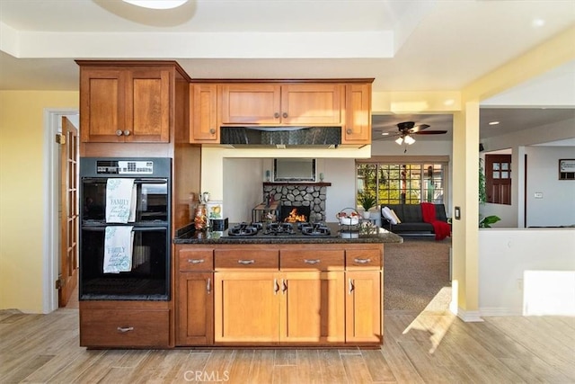 kitchen with a stone fireplace, dark stone counters, ceiling fan, black appliances, and light hardwood / wood-style flooring