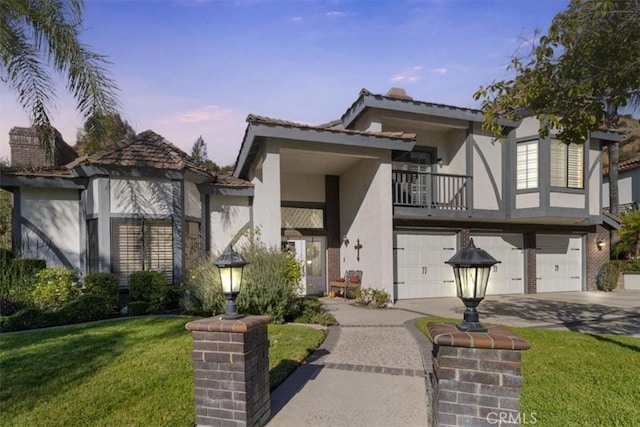 tudor-style house featuring a garage, concrete driveway, a balcony, a yard, and stucco siding