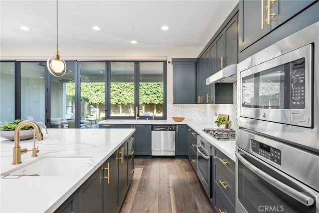 kitchen featuring light stone counters, under cabinet range hood, stainless steel appliances, a sink, and hanging light fixtures