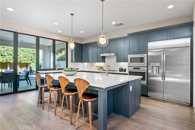 kitchen with tasteful backsplash, an island with sink, light stone counters, built in appliances, and pendant lighting