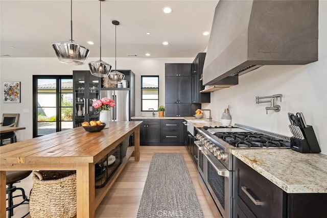 kitchen featuring light stone countertops, light wood-type flooring, wall chimney range hood, high quality appliances, and hanging light fixtures