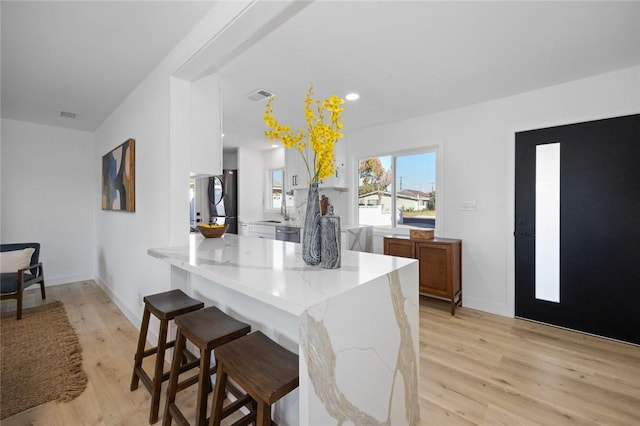 kitchen with kitchen peninsula, stainless steel fridge, a kitchen bar, light stone counters, and light hardwood / wood-style flooring