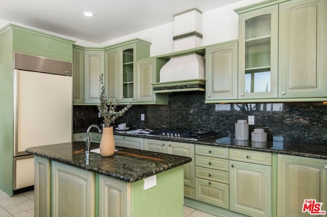 kitchen with custom exhaust hood, paneled built in refrigerator, dark stone countertops, tasteful backsplash, and stainless steel gas cooktop